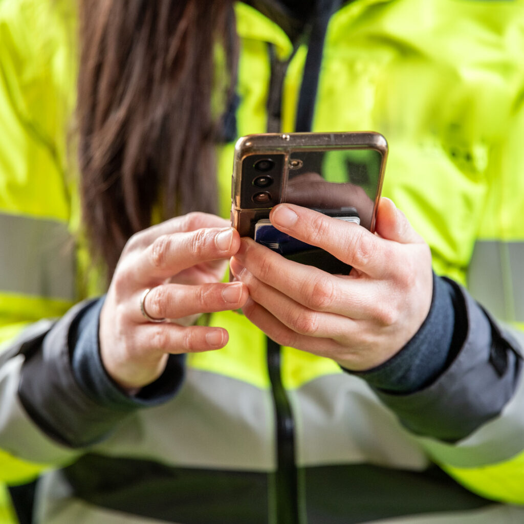 Illustrasjonsbilde: Hender med mobiltelefon og gul sikkerhetsjakke.